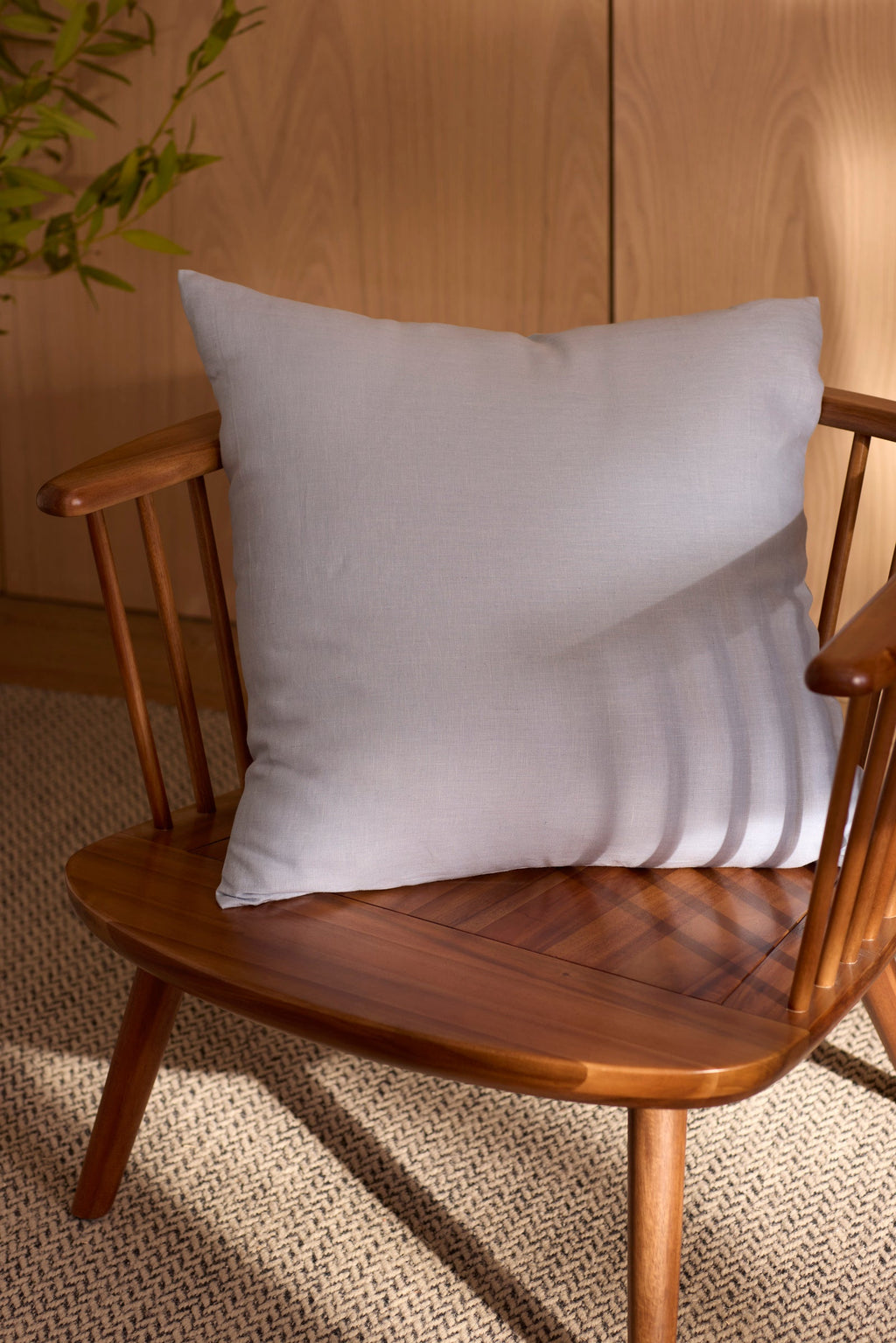 A Cozy Earth Linen Throw Pillow in light gray rests on a wooden spindle-back chair atop a textured beige rug, with sunlight and greenery in the background. |Color:Breeze|Size:24" x 24"
