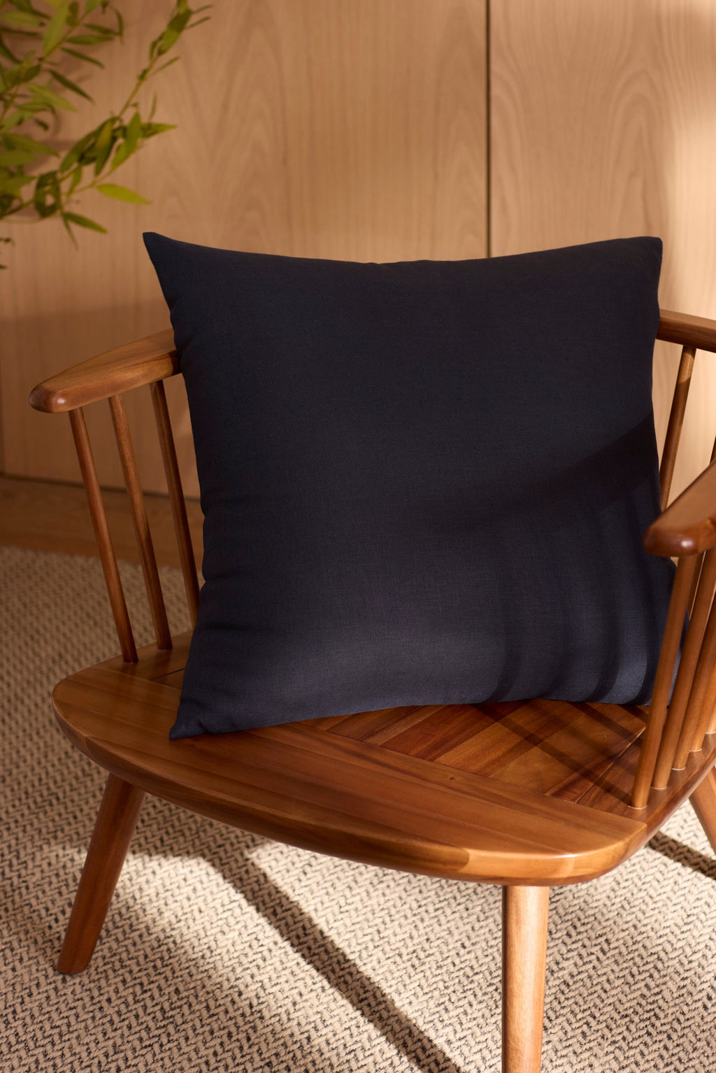 The Cozy Earth Linen Throw Pillow in navy blue sits on a wooden spindle-back chair atop a textured beige rug, with a green plant and light wood paneling in the background.|Color:Eclipse|Size:26" x 26"