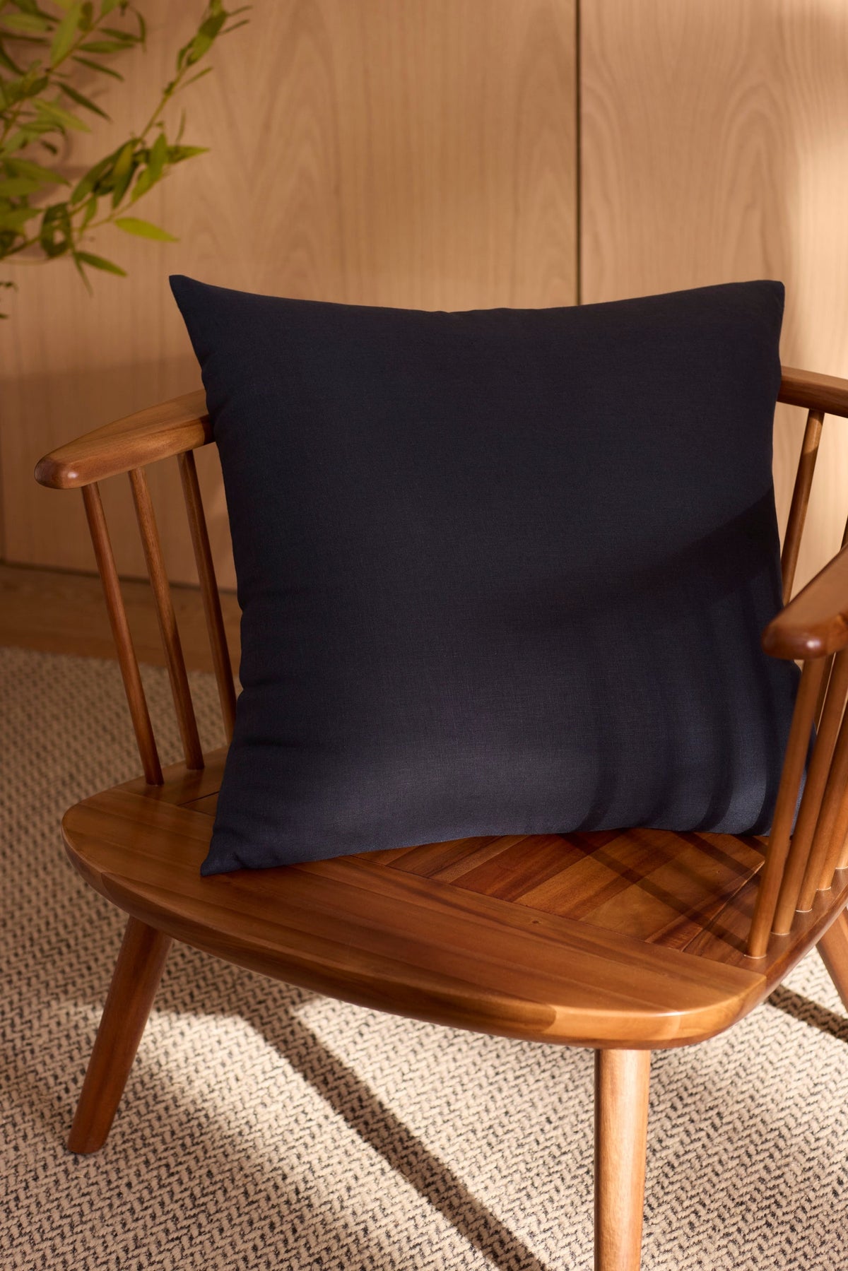 The Cozy Earth Linen Throw Pillow in navy blue sits on a wooden spindle-back chair atop a textured beige rug, with a green plant and light wood paneling in the background.|Color:Eclipse|Size:26" x 26"