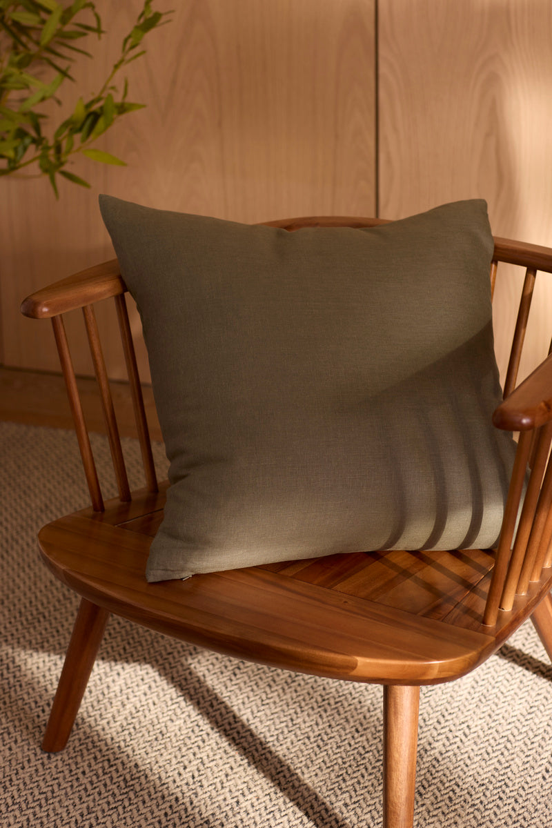 A wooden chair with spindle arms holds the Cozy Earth Linen Throw Pillow in olive green. Soft sunlight casts shadows on the pillow and chair, with a green plant and light wood paneling visible in the background.|Color:Olive |Size:20" x 20"