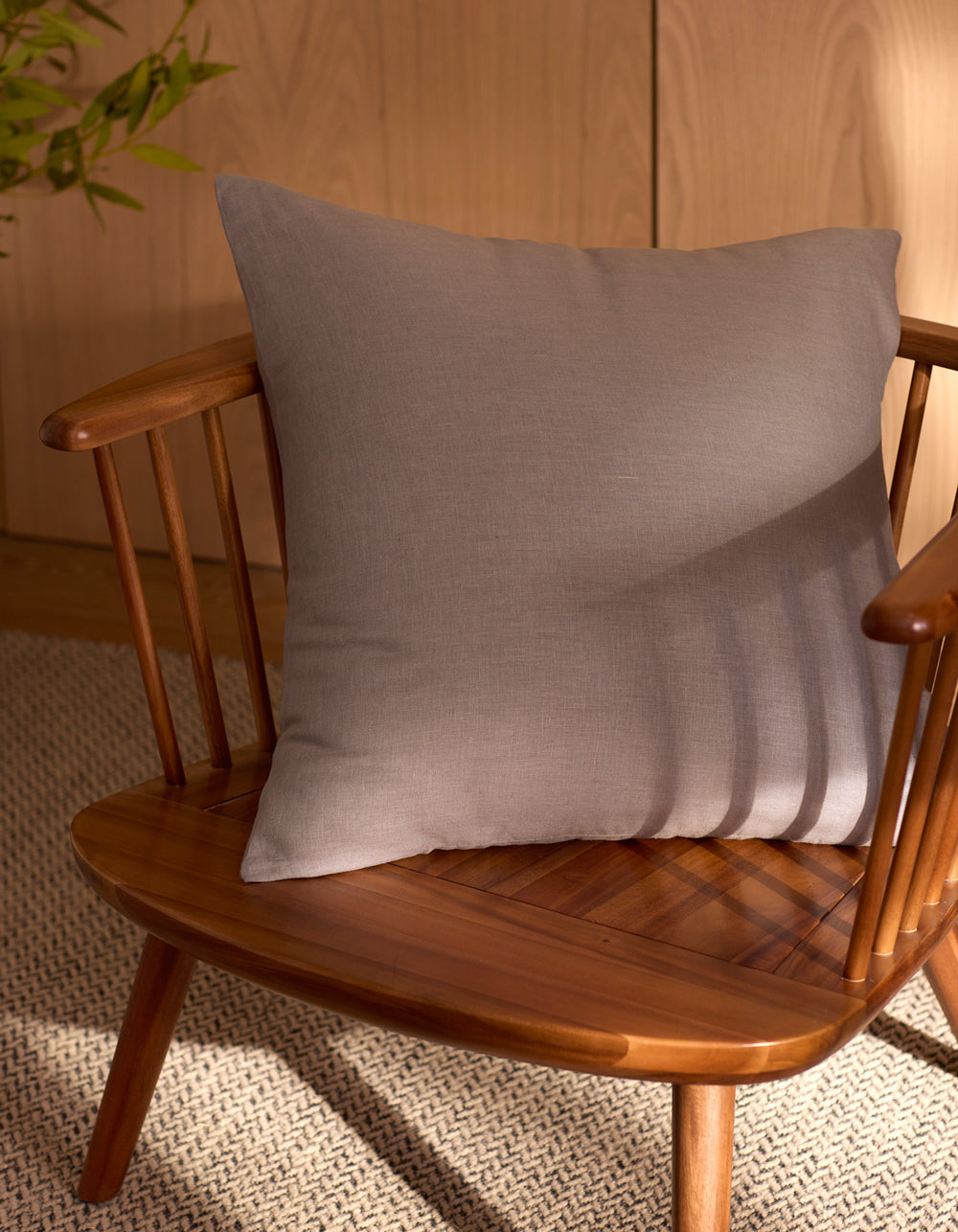 The Cozy Earth Linen Throw Pillow in light gray rests on a wooden spindle-back chair. Sunlight creates soft shadows, while a woven rug and green plant enhance the inviting background.|Color:Steel |Size:20" x 20"