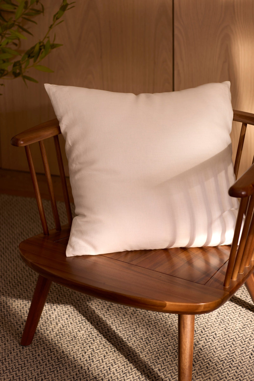 A Cozy Earth Linen Throw Pillow sits on a wooden chair with vertical slats, placed on a textured rug. Sunlight highlights the chair, with a wooden cabinet and green plant nearby. |Color:White |Size:24" x 24"