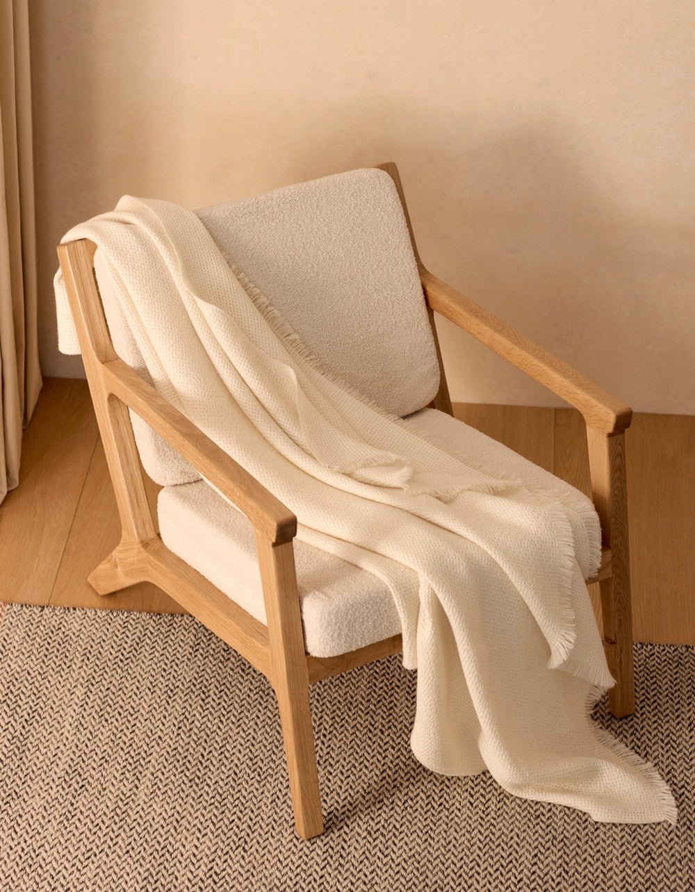 A modern wooden armchair with light beige upholstery sits on a herringbone rug, draped with the Cozy Earth Alpaca Waffle Throw. The soft blanket and minimal setting create a warm, inviting atmosphere.|Color:Creme