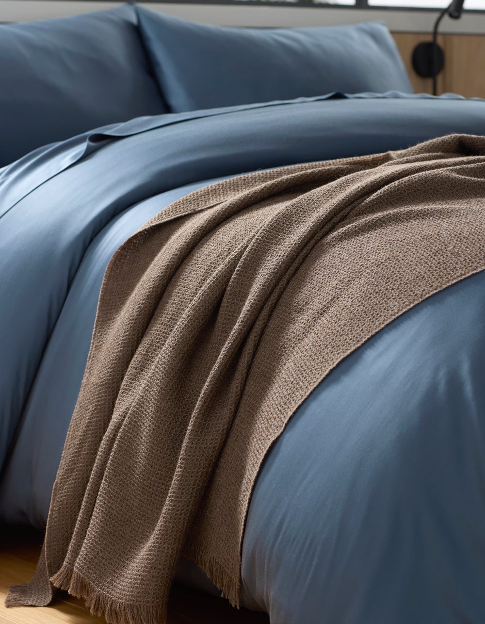 A close-up of a neatly made bed with smooth blue bedding and a Cozy Earth Alpaca Waffle Throw in brown draped casually over the edge.|Color:Ebony