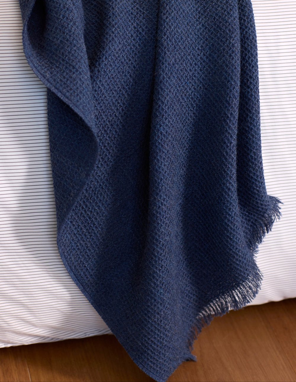 The Alpaca Waffle Throw by Cozy Earth, a textured dark blue blanket with fringed edges, drapes over a bed with white and gray striped sheets on a wooden floor.|Color:Slate Blue