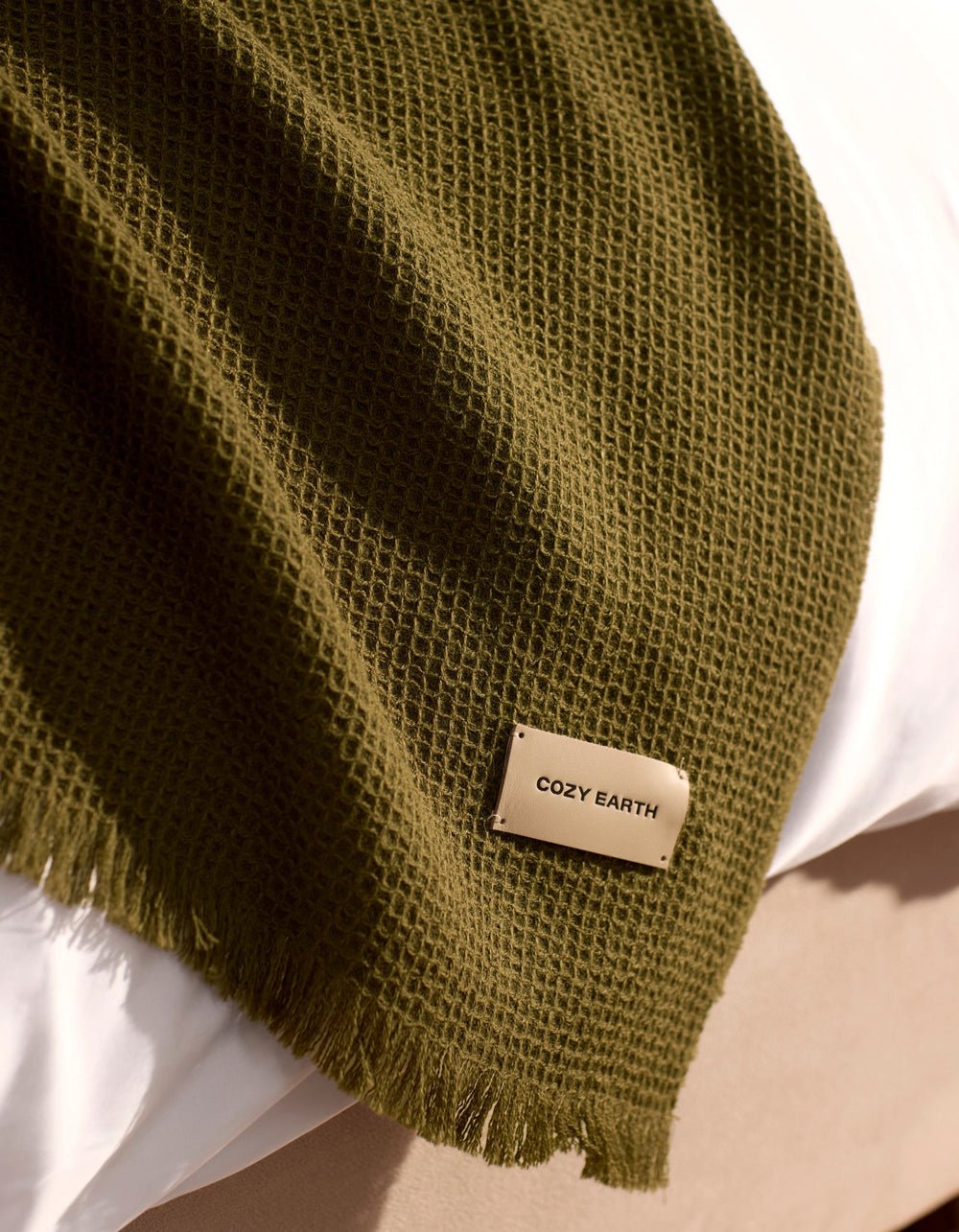 A Cozy Earth Alpaca Waffle Throw in dark green, featuring a textured waffle weave and fringed edges, is draped over a white surface. A beige tag on the throw displays the brand name "Cozy Earth.|Color:Utility Green
