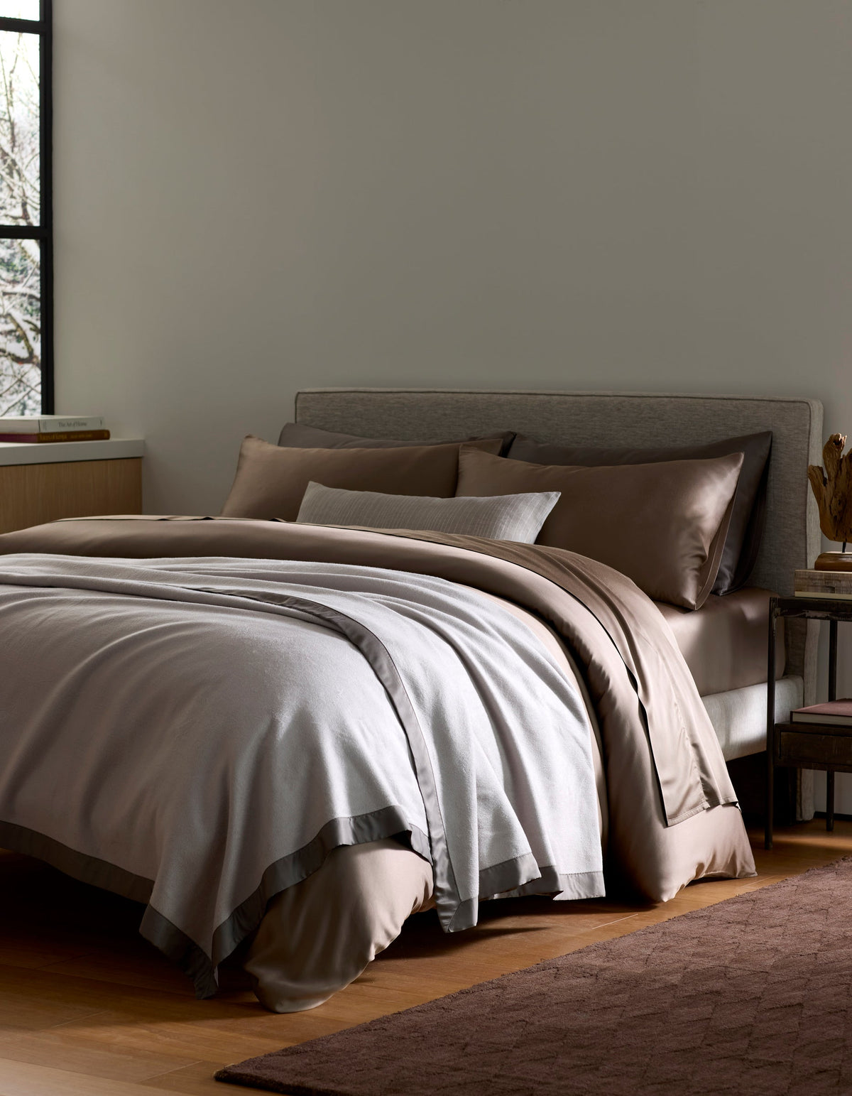 A neatly made bed features the Cozy Earth Bamboo Blanket in brown and light gray, with pillows against a gray upholstered headboard beside a window and wooden nightstand on hardwood floors.|Color:Dove Grey/Charcoal