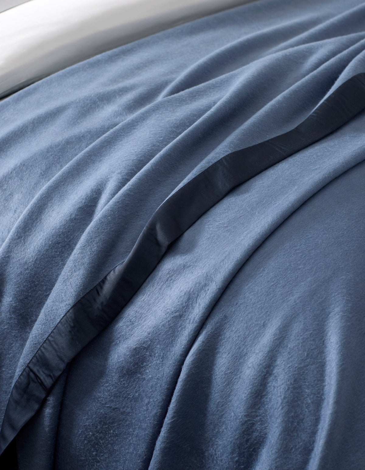 A close-up of a bed featuring the Cozy Earth Bamboo Blanket, showcasing its soft blue velvet-like texture, smooth folds, and a satin edge trim.|Color:Pacific Blue/Navy