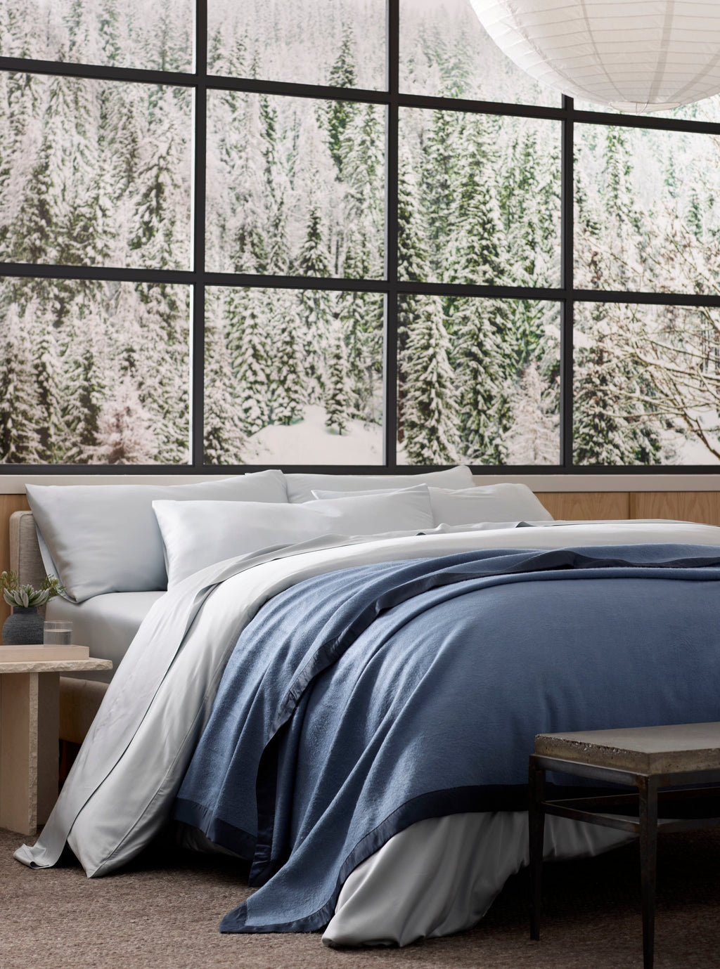 A neatly made bed, topped with a Cozy Earth Bamboo Blanket, sits before large windows revealing a snowy evergreen forest. A small table and bench are nearby, while a paper lantern hangs overhead.|Color:Pacific Blue/Navy