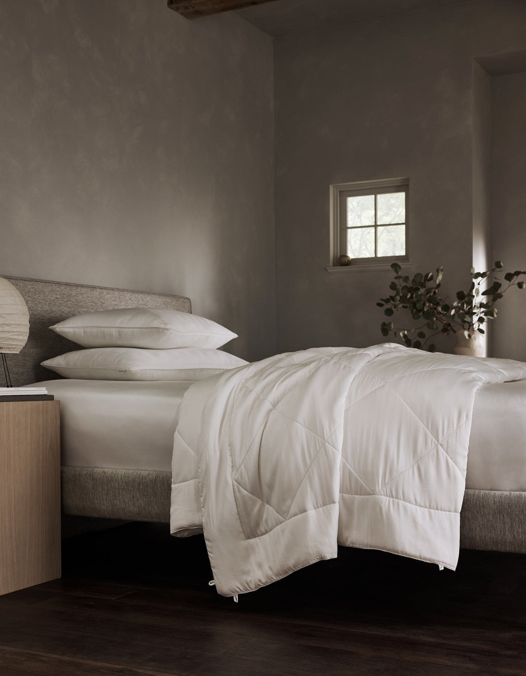 A bed dressed in Cozy Earth's Bamboo Viscose Comforter with white pillows anchors a minimalist bedroom featuring gray walls, a small window, a nightstand with lamp, and a vase of greenery.|Filling:Extra