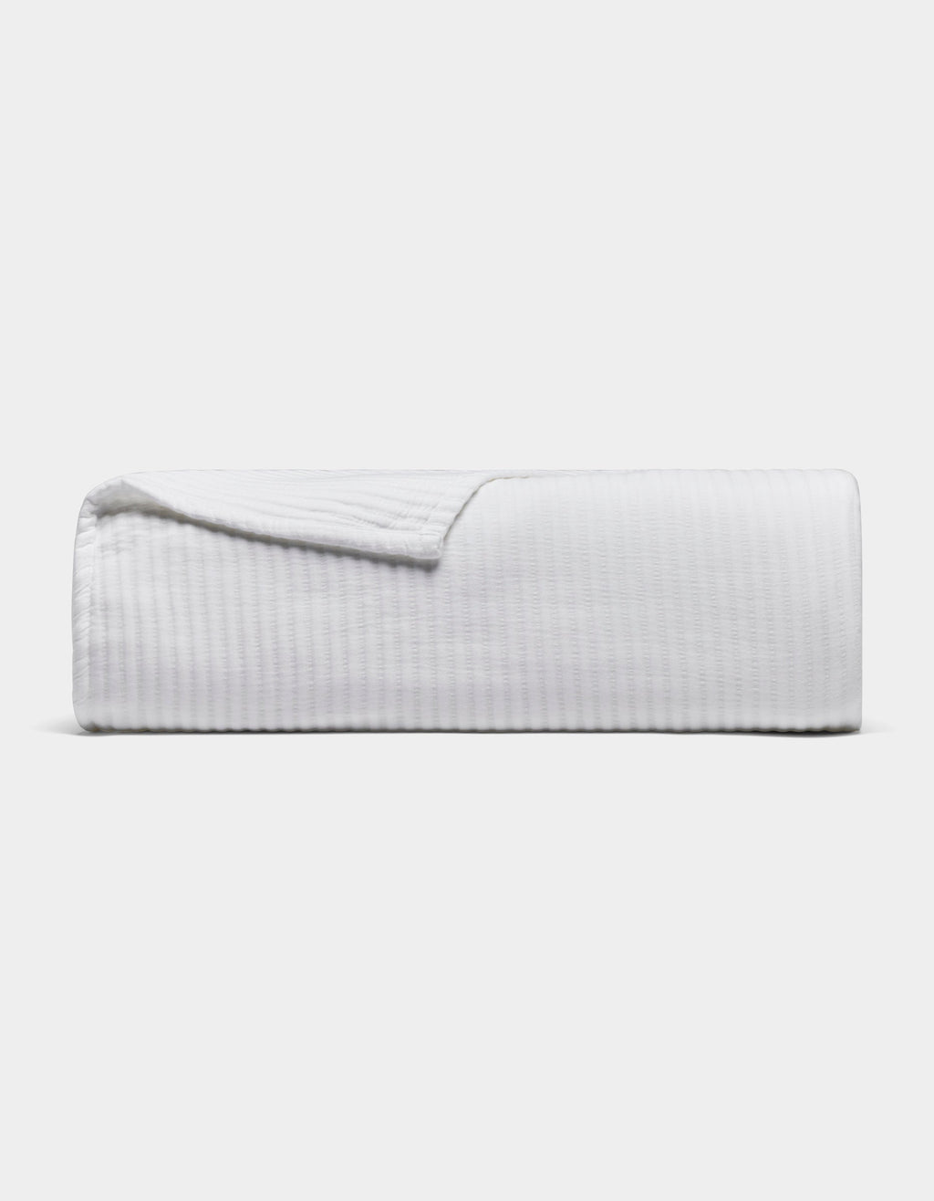 The Cozy Earth Bamboo Coverlet, a white textured blanket, is neatly folded with one corner slightly lifted, set against a plain light gray background.|Color:White