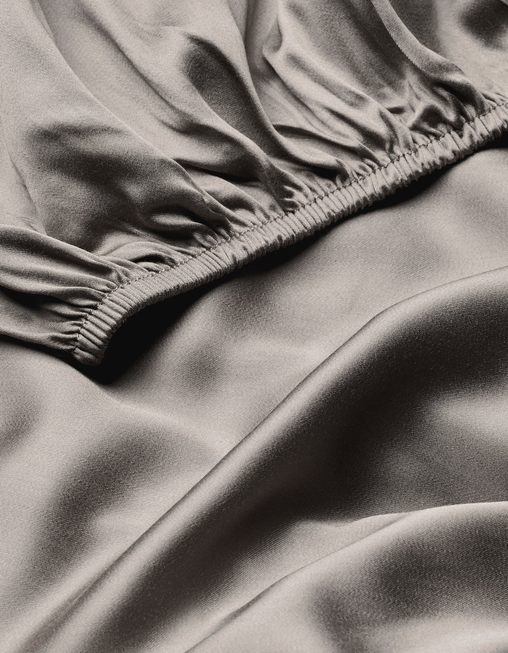 A close-up of the Cozy Earth Bamboo Fitted Sheet shows smooth, light gray satin fabric with gentle folds and a soft, shiny surface. An elastic gathered seam runs diagonally across the image.|Color:Charcoal