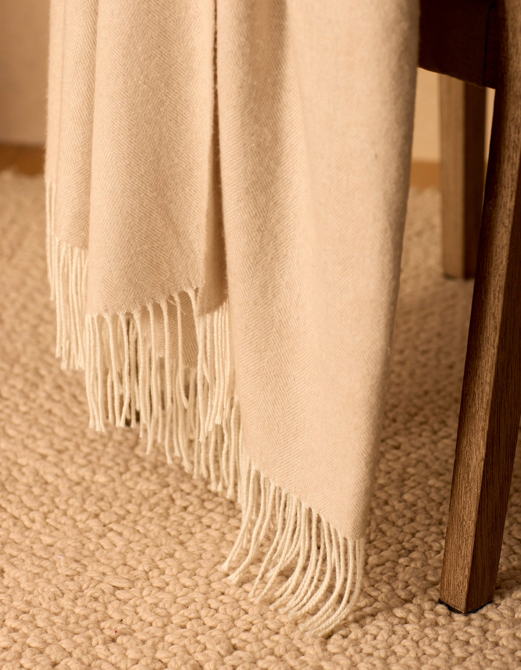The Bamboo Herringbone Tassel Throw by Cozy Earth, in a cream color with fringe, is draped over a wooden chair on a textured beige carpet.|Color:Oatmeal