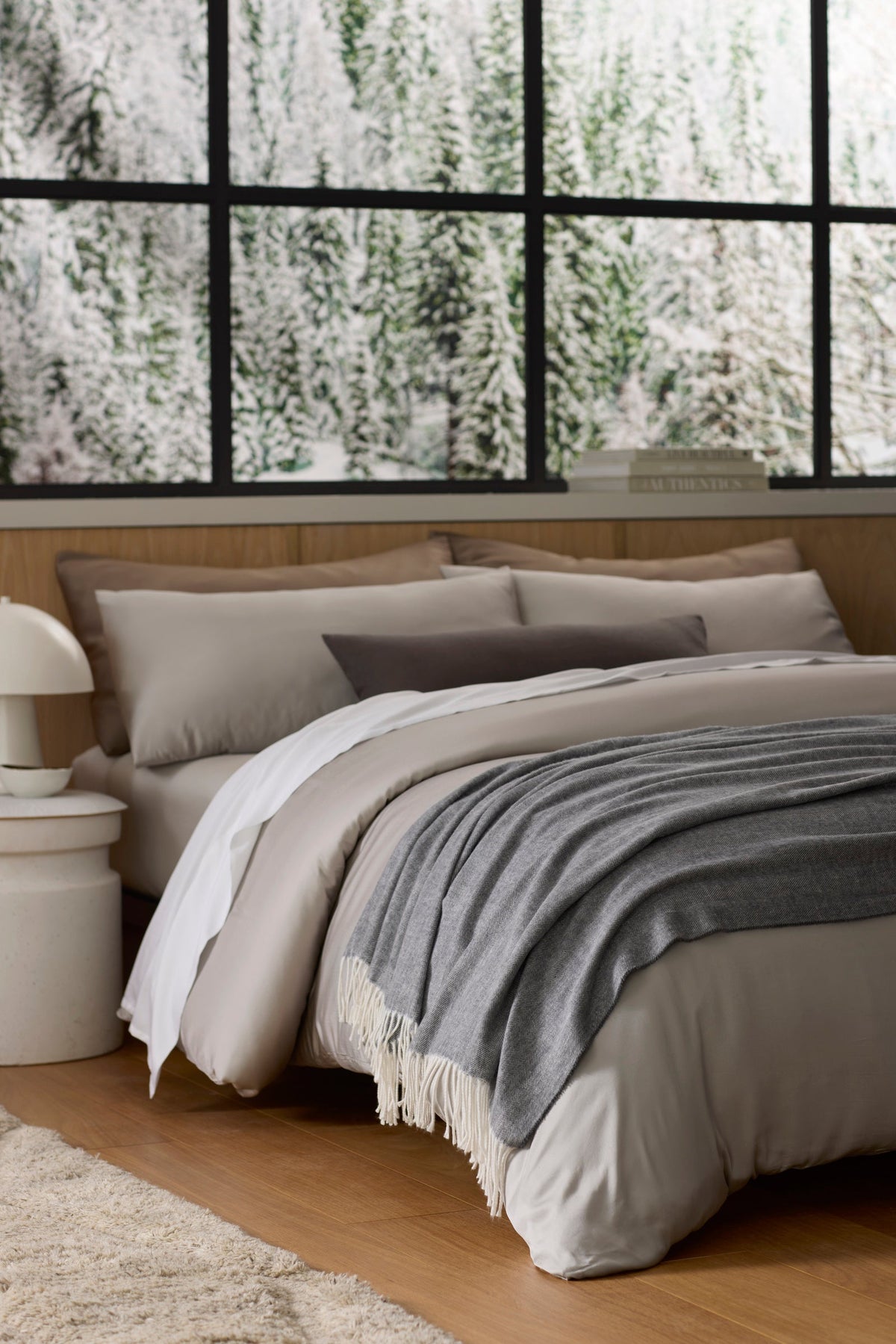 A cozy bed with gray and beige bedding sits by a large window overlooking a snowy forest. A Bamboo Herringbone Tassel Throw by Cozy Earth is draped over the bed, and a white lamp rests on the bedside table, creating a warm, inviting atmosphere.|Color:Shadow