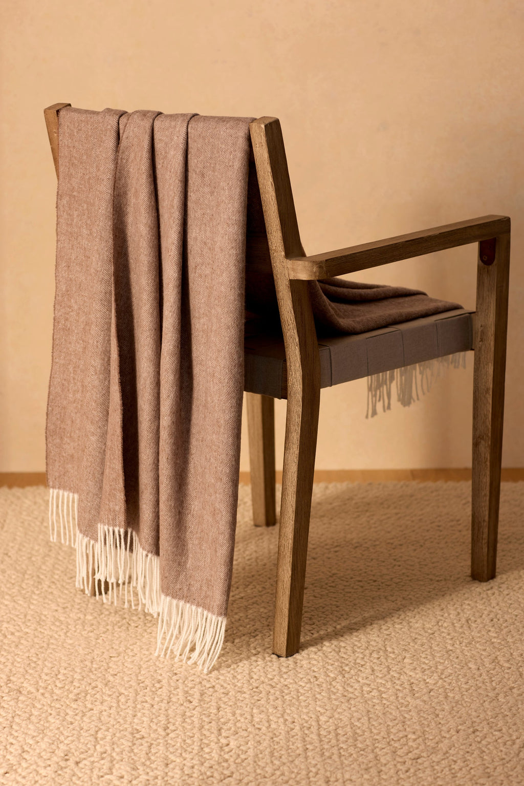 A wooden chair with the Cozy Earth Bamboo Herringbone Tassel Throw in taupe, featuring white fringed ends, is draped over the backrest and sits on a textured beige carpet against a light tan wall.|Color:Truffle