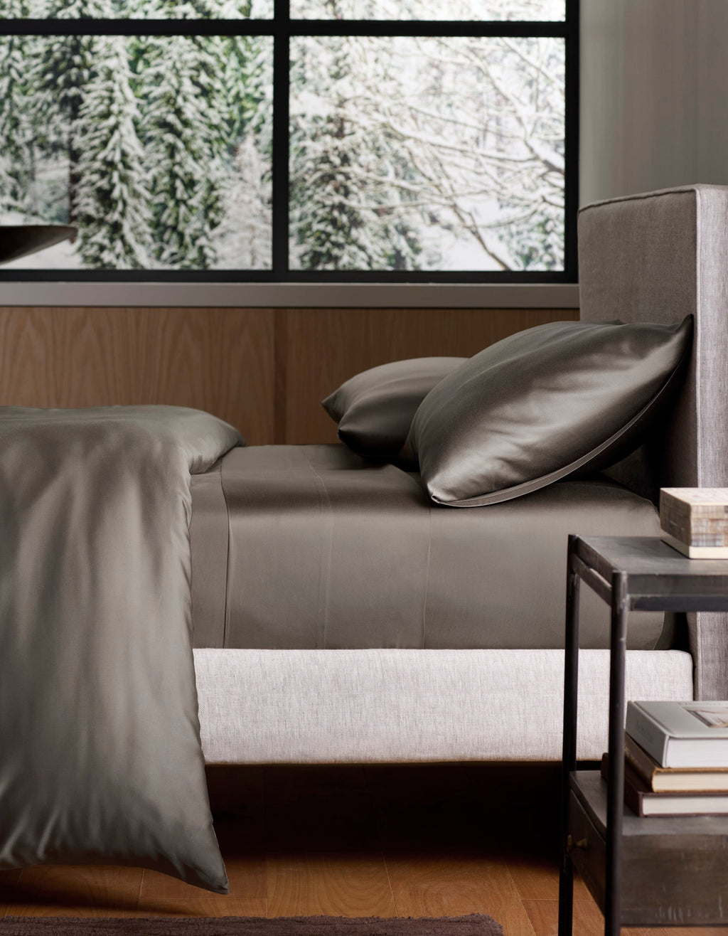 A neatly made bed with Cozy Earth’s Bamboo Sheet Set in dark gray rests by a modern nightstand. Behind the bed, a large window reveals a snowy forest with pine trees. |Color:Charcoal