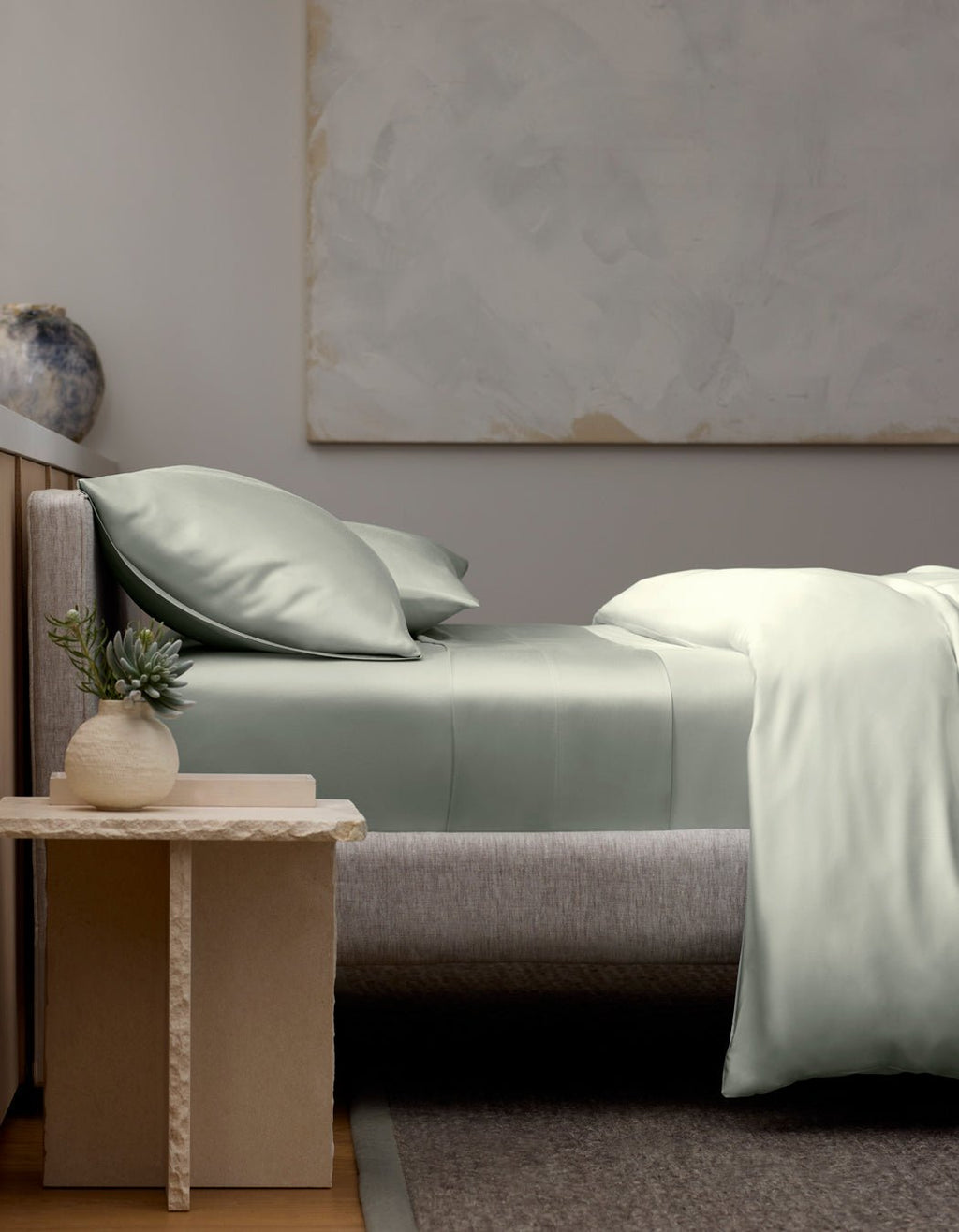 A modern bedroom featuring a neatly made bed with Cozy Earth’s Bamboo Fitted Sheet, two pillows, and a gray upholstered headboard. A small table with a potted succulent and ceramic vase sits beside the bed. Abstract art hangs on the wall.|Color:Laurel