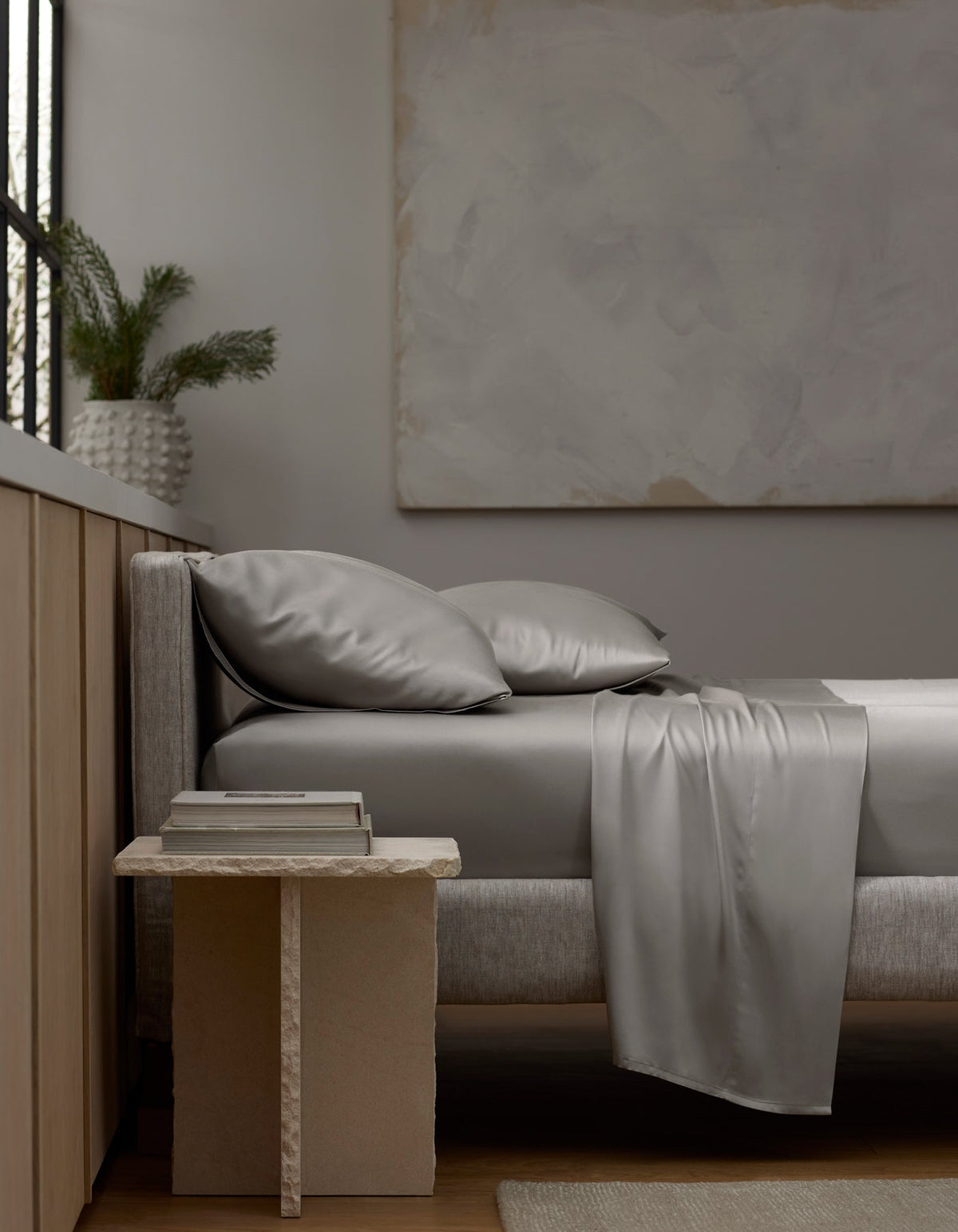 A modern bedroom with a neatly made bed featuring Cozy Earth’s Bamboo Sheet Set in shiny silver-gray. A textured side table holds stacked books, and a vase with greenery sits on the windowsill, creating minimalist neutral-toned decor. |Color:Light Grey