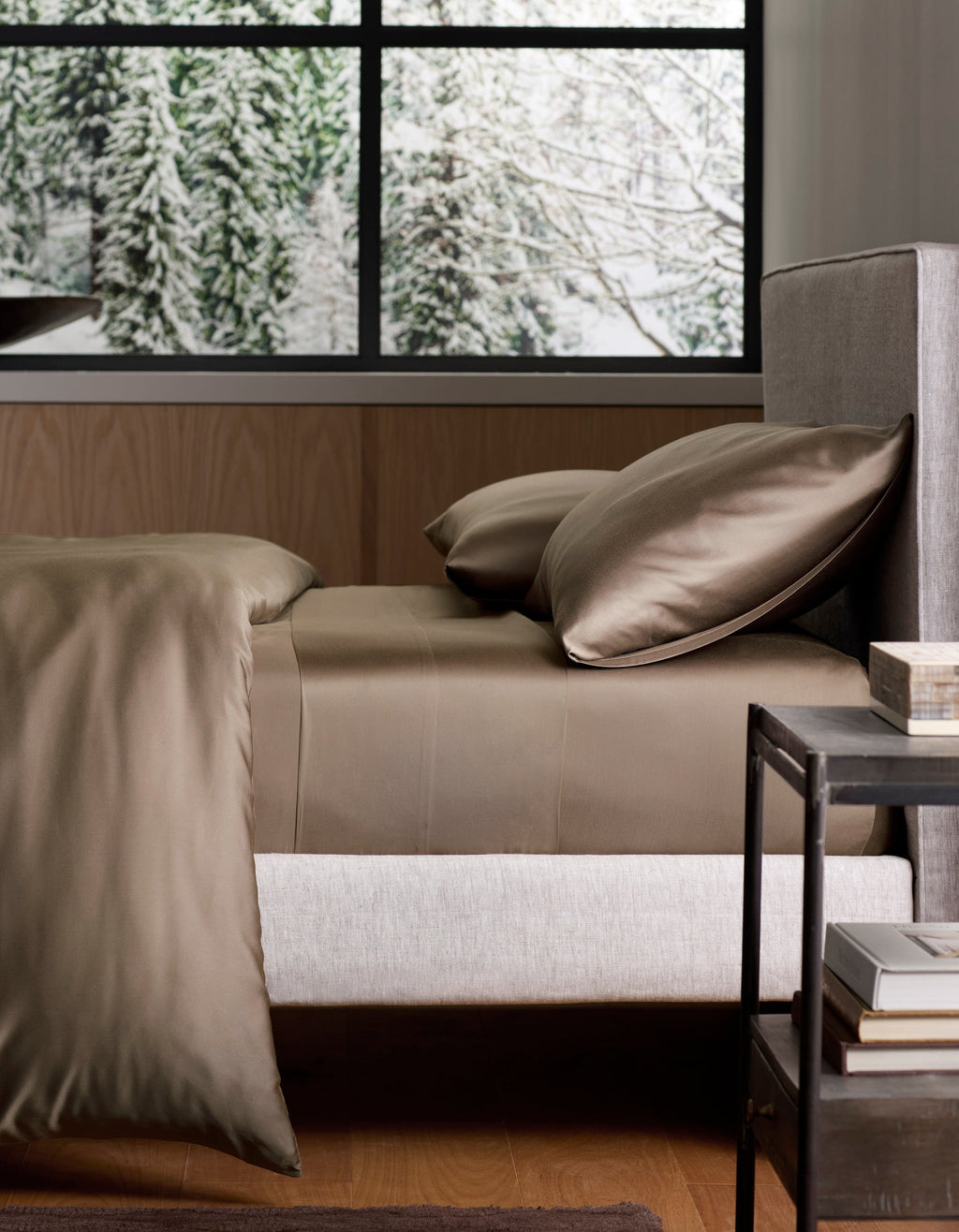 A neatly made bed with Cozy Earth Bamboo Sheet Set in silky brown lies next to a black nightstand, framed by a large window revealing snow-covered trees outside. |Color:Walnut