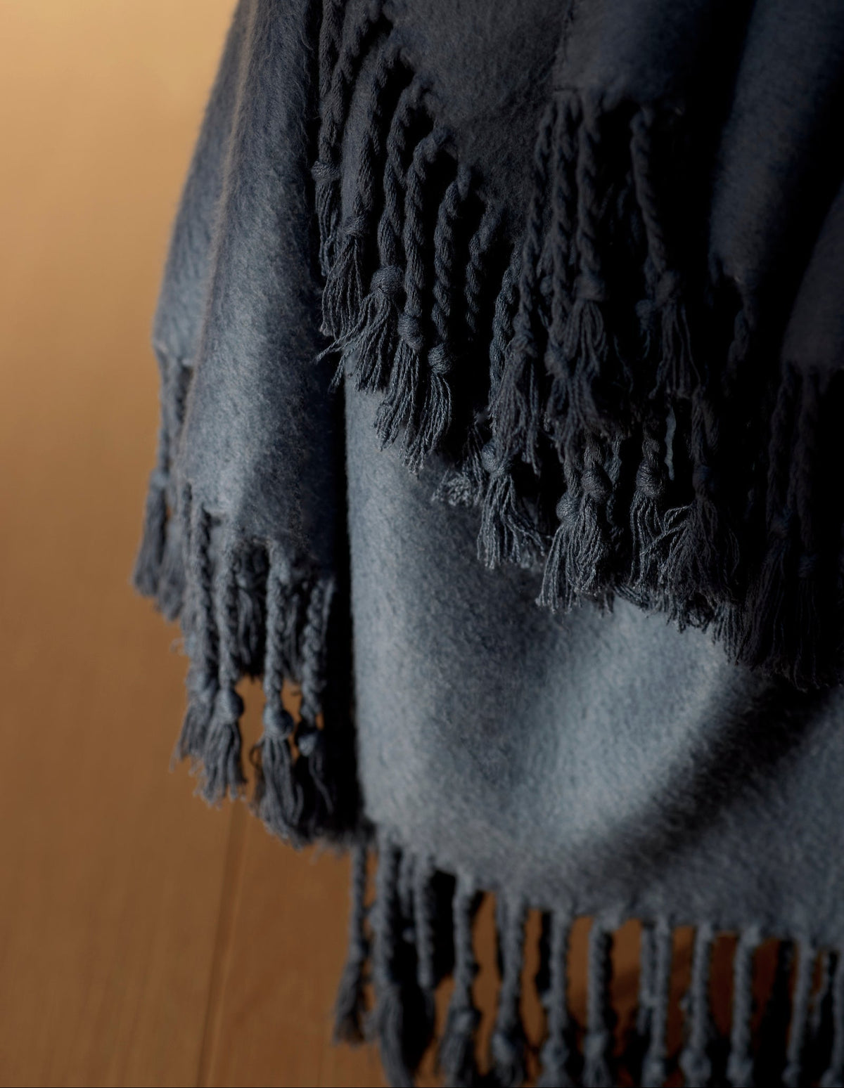 A close-up of the Cozy Earth Bamboo Tassel Throw, a dark blue blanket with fringe, draped over the edge of a wooden surface.|Color:Pacific Blue