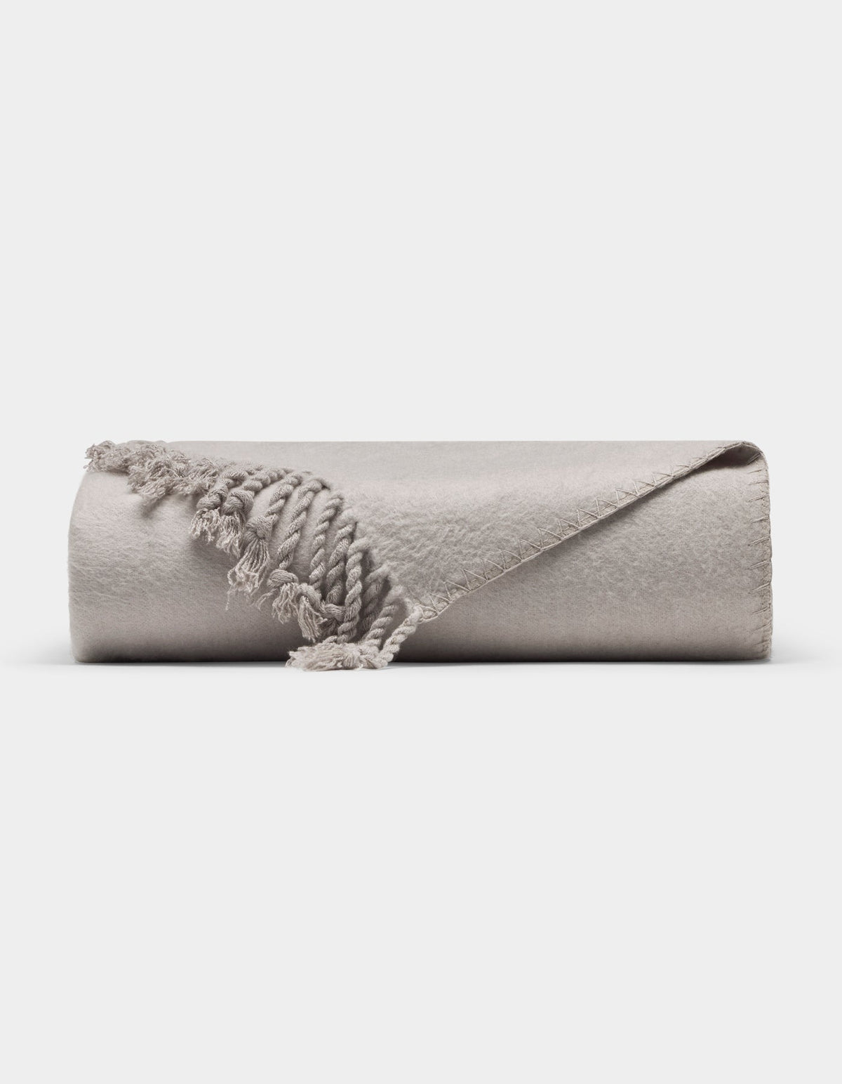 The Cozy Earth Bamboo Tassel Throw is a neatly rolled, light gray blanket with fringed tassels, shown on a white background. One corner is flipped to reveal its soft texture and detailed edging.|Color:Dove Grey