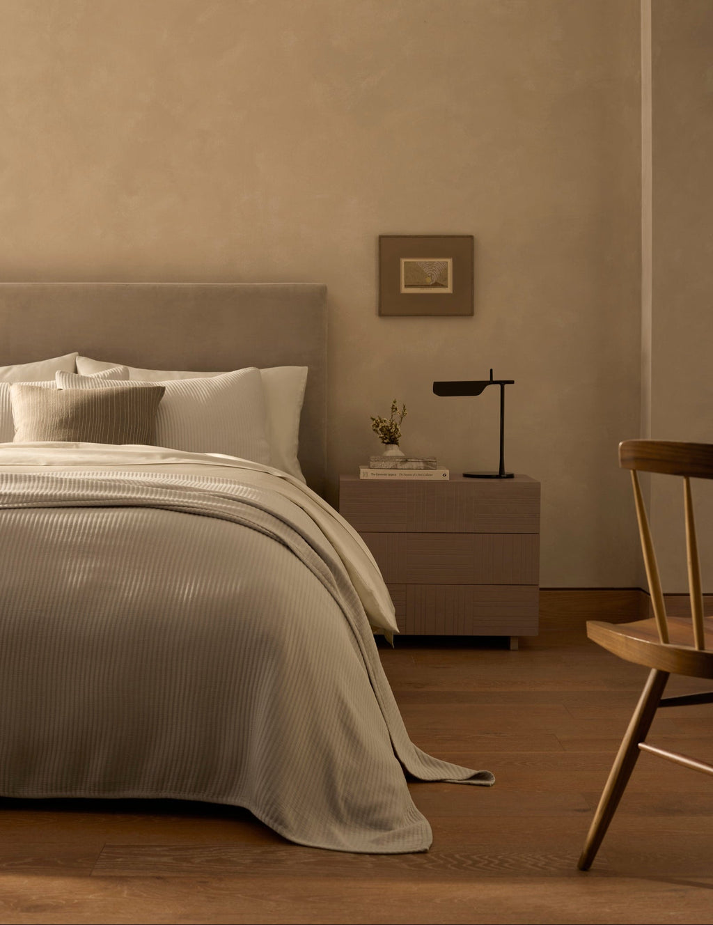 A minimalist bedroom features a neatly made bed with the Cozy Earth Bamboo Coverlet in beige tones, a wooden nightstand topped with a modern black lamp and small vase, a framed picture on the wall, and a wooden chair on hardwood floors.|Color:Driftwood