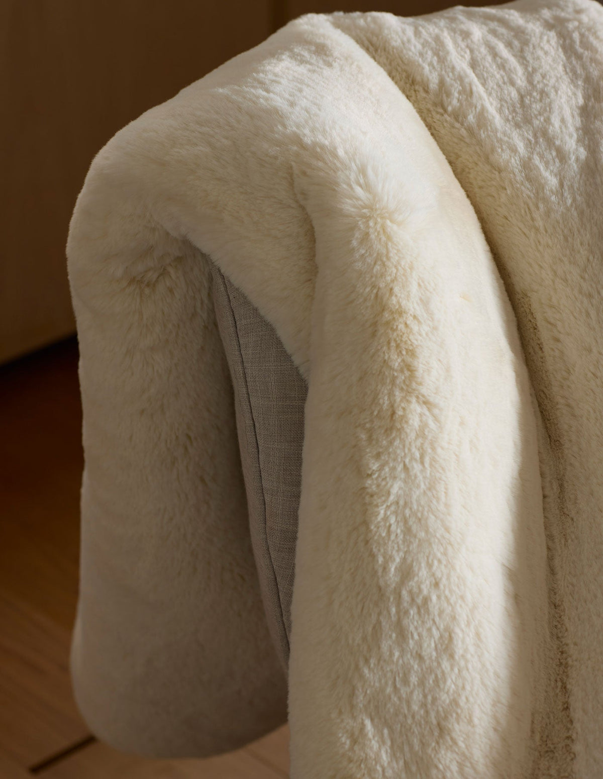 The Cozy Earth Cuddle Blanket, in soft cream-colored faux fur, is draped over a light gray chair, its cozy texture highlighted by natural light.|Color:Creme