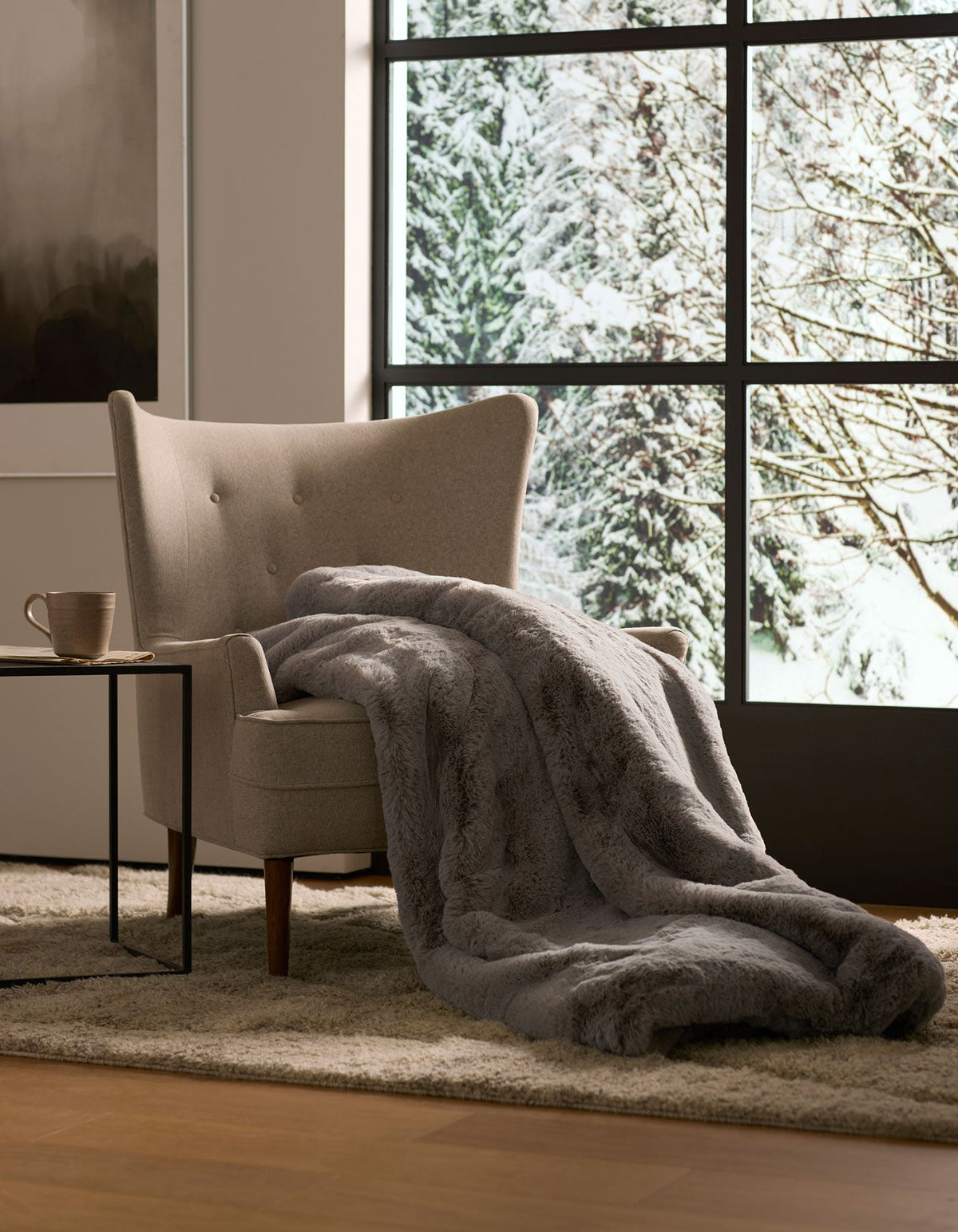 A gray armchair with a Cozy Earth Cuddle Blanket draped over it sits by a large window overlooking snowy trees, while a mug rests on the side table nearby.|Color:Dove Grey