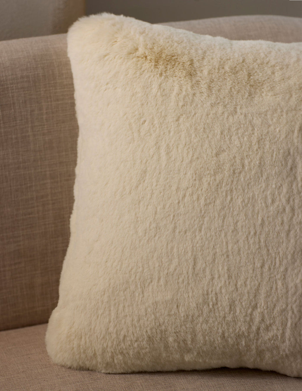 A close-up of the Cuddle Pillow by Cozy Earth, featuring a fluffy cream color and soft texture, placed on a light beige fabric sofa.|Color:Creme |Size:24" x 24"