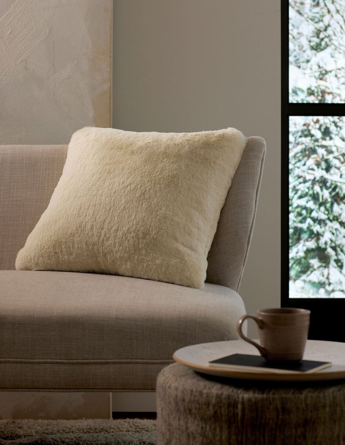 A cozy interior scene with a light beige sofa, a fluffy Cozy Earth Cuddle Pillow in cream, a round ottoman holding a cup and saucer, and a window framing a snowy outdoor view.|Color:Creme |Size:24" x 24"