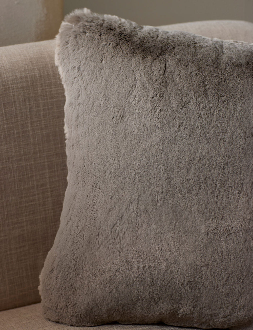 The Cozy Earth Cuddle Pillow, a large square in light gray faux fur, rests against the back of a beige fabric sofa.|Color:Dove Grey |Size:20" x 20"