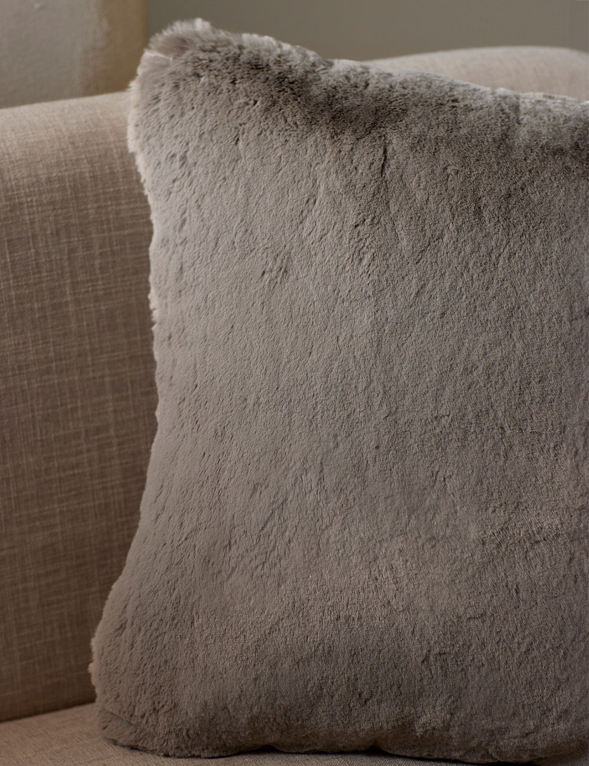 The Cozy Earth Cuddle Pillow, a large square in light gray faux fur, rests against the back of a beige fabric sofa.|Color:Dove Grey |Size:20" x 20"