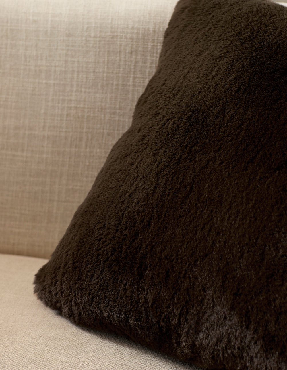 A Cozy Earth Cuddle Pillow in dark brown rests against the back of a light beige, textured fabric sofa.|Color:Espresso |Size:Euro
