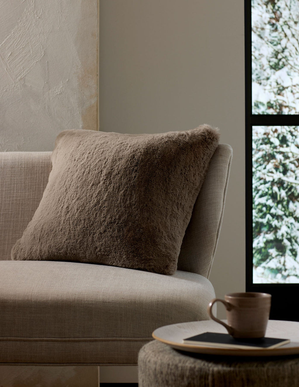 A cozy, neutral-toned living space features the Cozy Earth Cuddle Pillow in taupe on a beige sofa next to a round side table with a ceramic mug and book, framed by a window view of snowy trees.|Color:Walnut |Size:24" x 24"