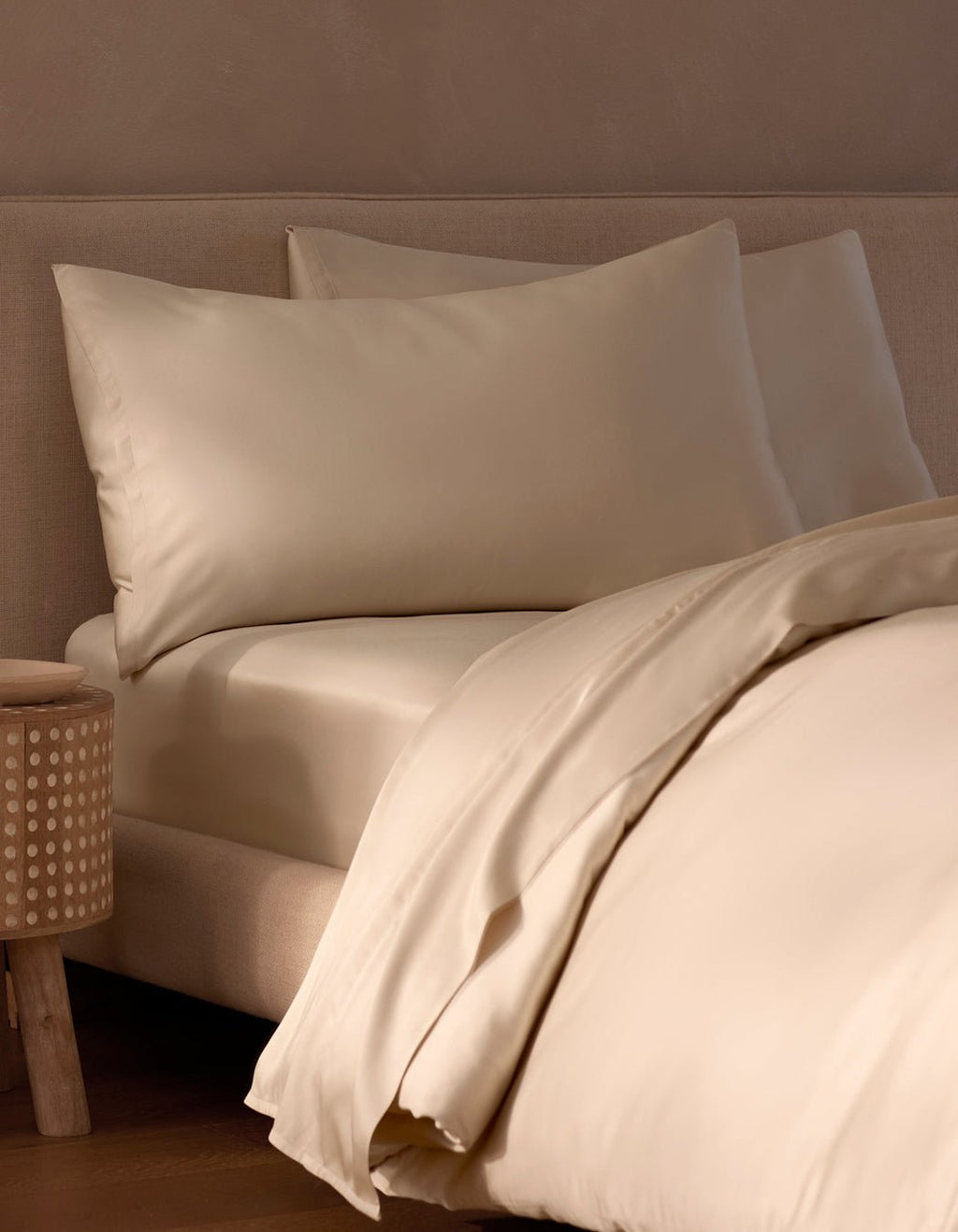 A cozy, minimalistic bedroom features a neatly made bed with cream-colored Cozy Earth Bamboo Pillowcases, sheets, and duvet, placed beside a wooden nightstand with a dotted pattern.|Color:Creme