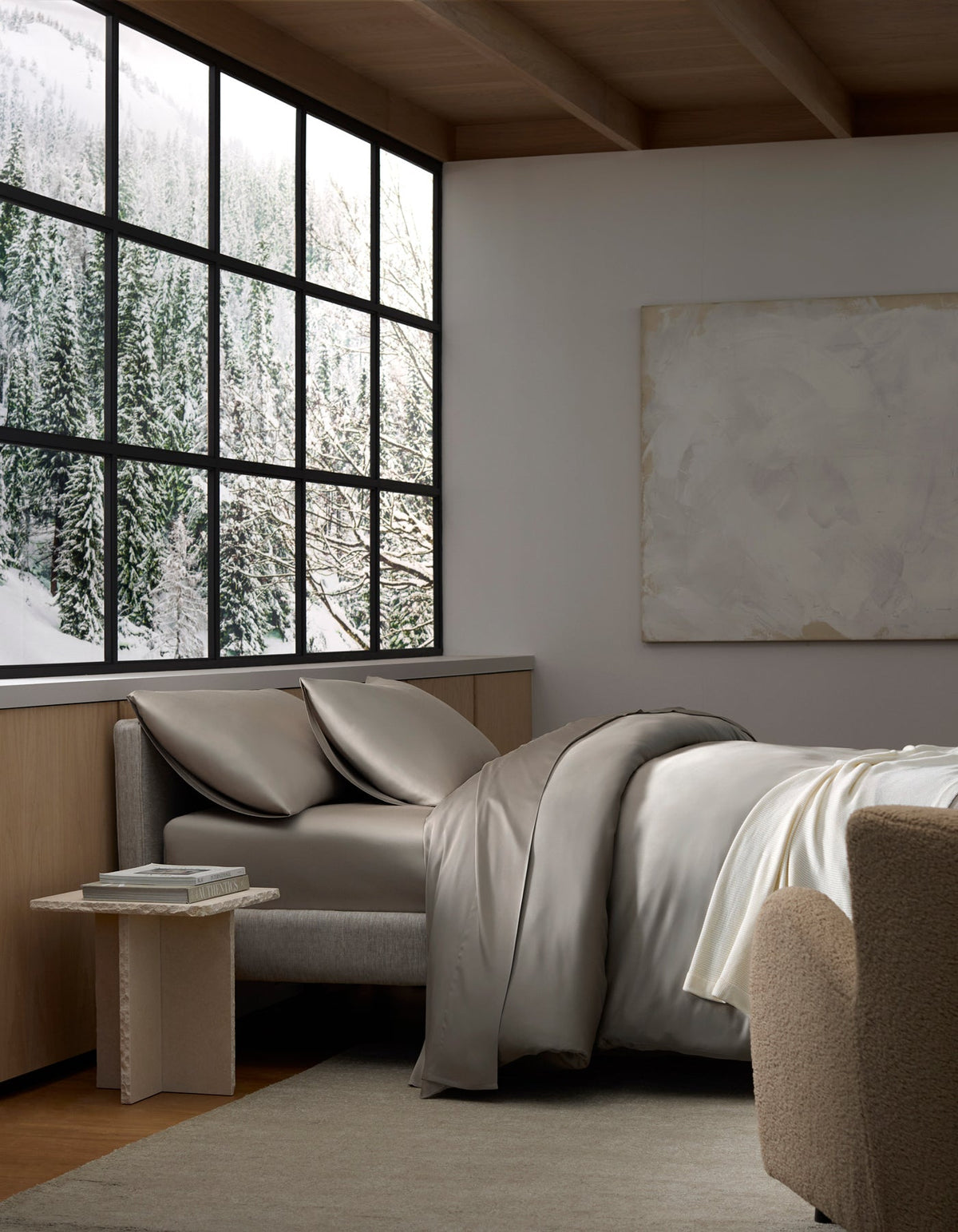 A cozy bedroom featuring Cozy Earth's Bamboo Bedding Deluxe Bundle, a gray bed and chair by the window with snowy forest views. Neutral tones, a soft rug, and a minimalistic painting create a serene, modern vibe.|Color:Dove Grey