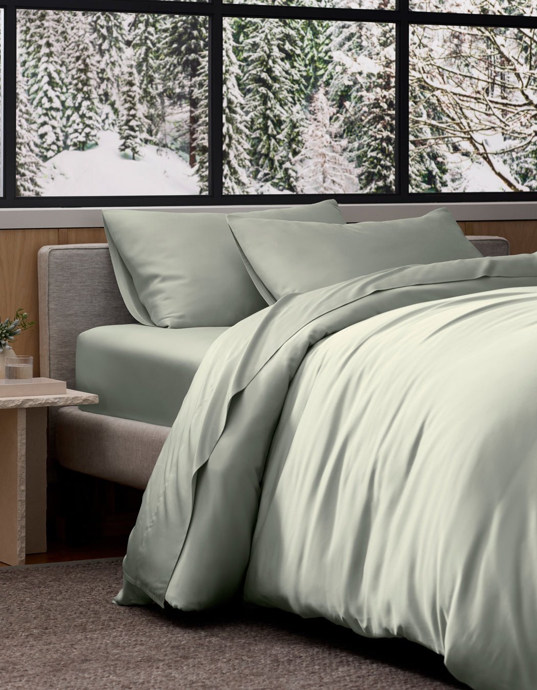 A neatly made bed features Cozy Earth's Bamboo Bedding Deluxe Bundle in light sage green, positioned by a window overlooking a snowy evergreen forest, creating a modern and cozy ambiance.|Color:Laurel