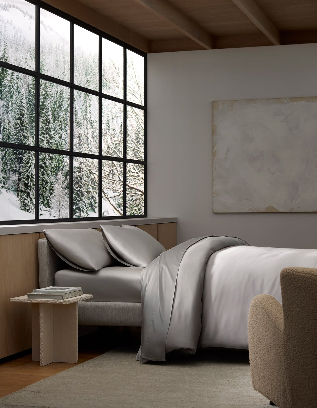 A cozy bedroom filled with natural light features the Cozy Earth Bamboo Bedding Deluxe Bundle on the bed, gray sheets and pillows, a small table, a beige chair, a large window overlooking a snowy forest, and a blank canvas on the wall.|Color:Light Grey