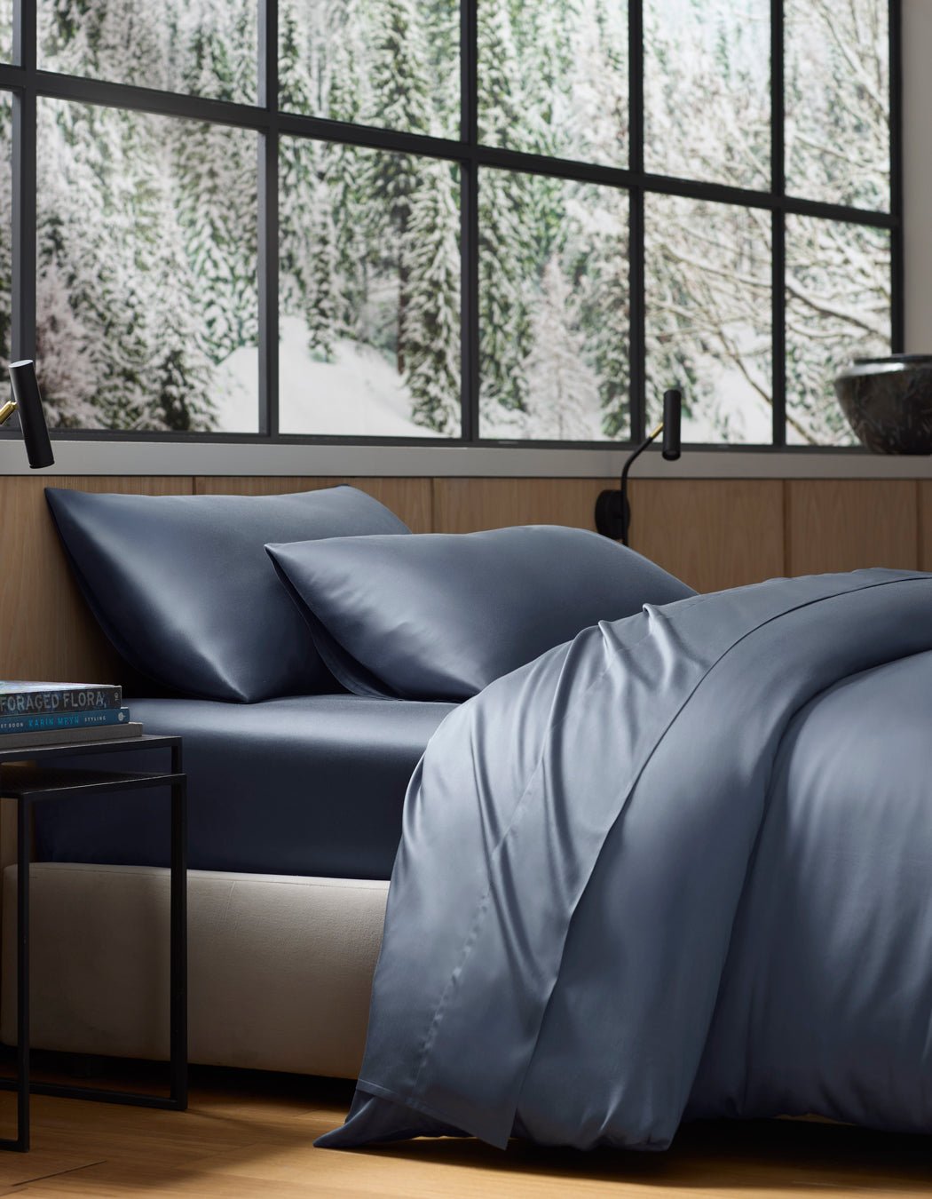 A neatly made bed featuring the Cozy Earth Bamboo Flat Sheet in dark blue sits by a large window overlooking a snowy forest. The room has wooden walls, a black lamp, and a small bedside table.|Color:Pacific Blue