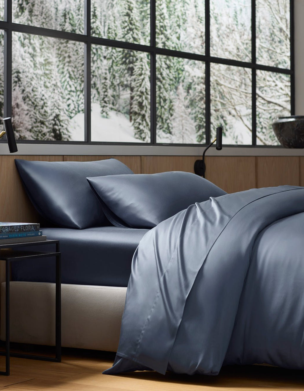 A neatly made bed featuring the Cozy Earth Bamboo Flat Sheet in dark blue sits by a large window overlooking a snowy forest. The room has wooden walls, a black lamp, and a small bedside table.|Color:Pacific Blue