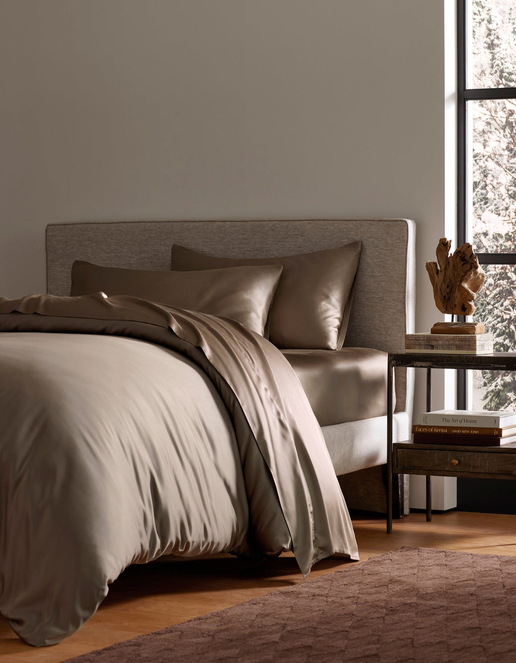A modern bedroom features a neatly made bed with Cozy Earth’s Bamboo Bedding Deluxe Bundle in taupe, paired with pillows. A wooden nightstand nearby holds books, a lamp, and decor, as sunlight adds warmth to the inviting space.|Color:Walnut