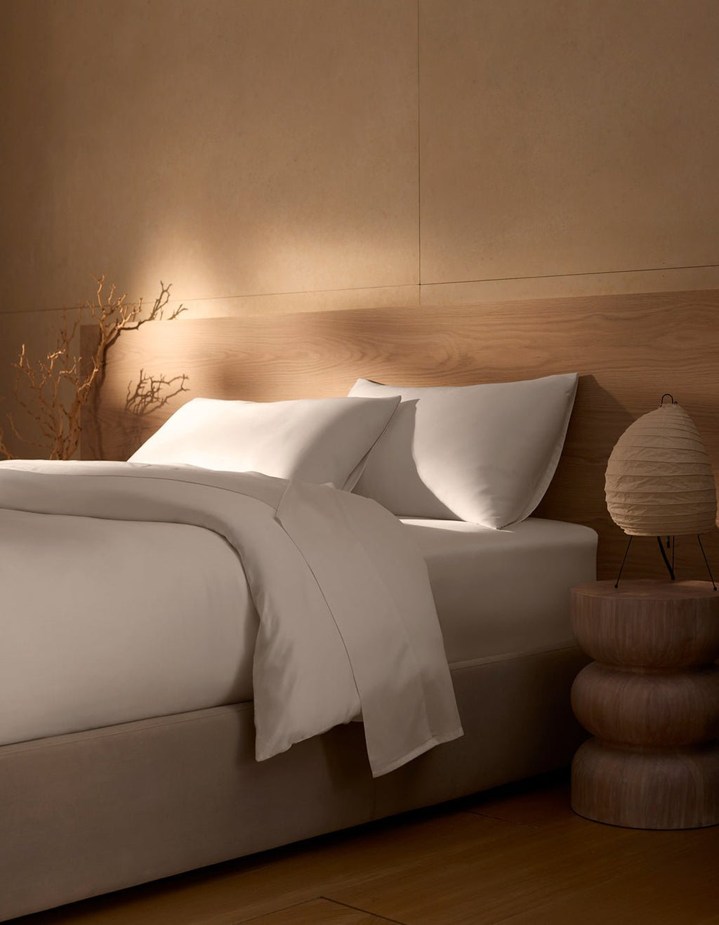 A neatly made bed featuring Cozy Earth's Bamboo Duvet Cover is set beside a wooden nightstand with a round lamp; beige walls, natural wood accents, and a dried branch in the corner create a calm, minimalist atmosphere.|Color:White
