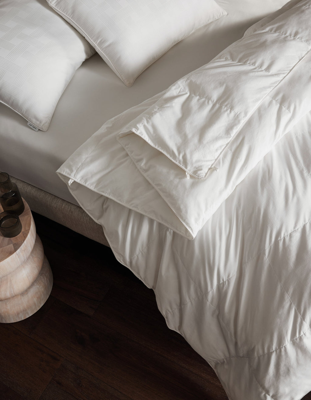 A neatly made bed features Cozy Earth's Down Alternative Comforter and white pillows beside a small wooden nightstand on a dark wood floor.|Filling:Lightweight
