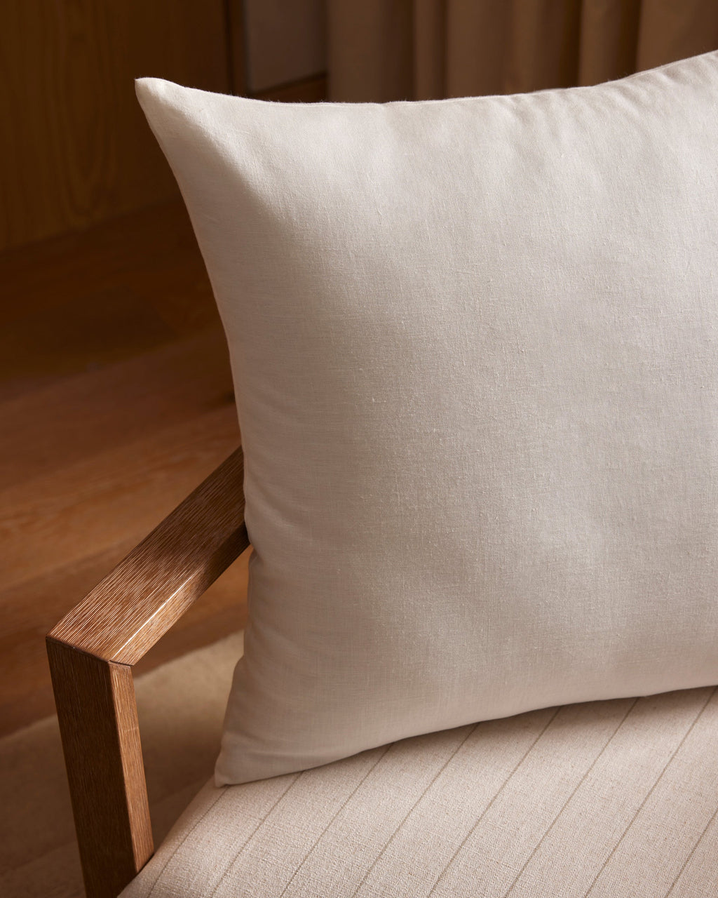 A close-up of Cozy Earth’s Linen Throw Pillow in light beige, placed on a wooden chair with a textured cushion. The scene is warm and minimalistic, featuring soft lighting and wooden flooring in the background.|Color:Birch |Size:24" x 24"