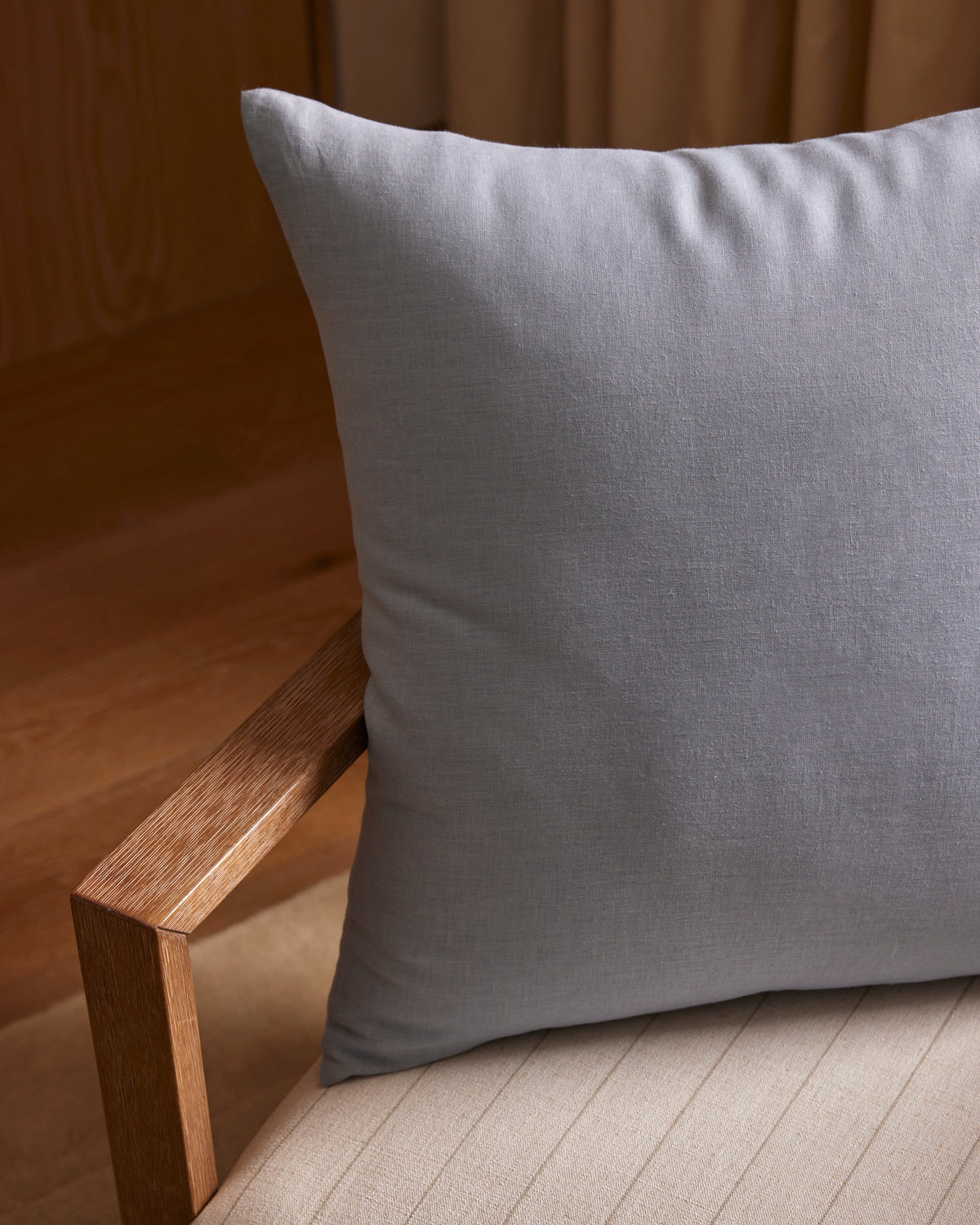 A Cozy Earth Linen Throw Pillow in light gray rests on a beige upholstered chair with wooden arms, set against wooden flooring and tan curtains.|Color:Breeze |Size:26" x 26"
