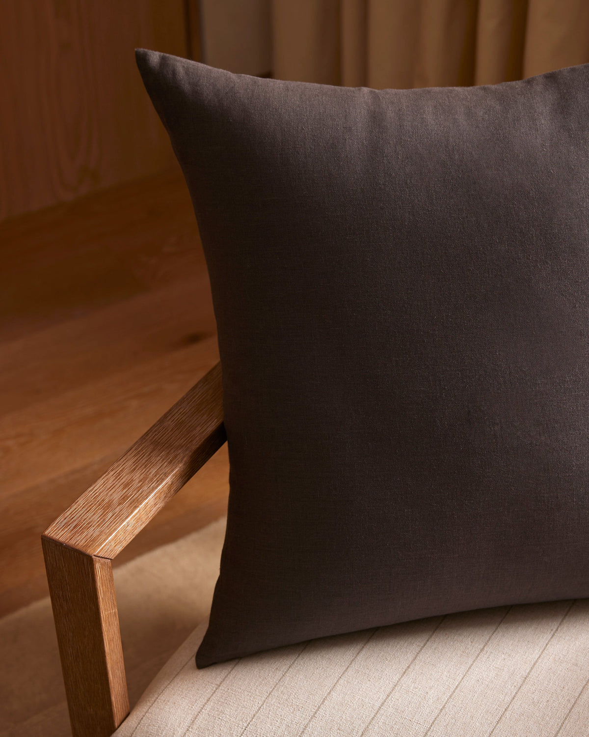 A close-up of the Cozy Earth Linen Throw Pillow in dark brown, placed on a light cushioned chair with a wooden armrest, set in a softly lit room featuring wooden flooring and beige curtains.|Color:Coal|Size:24" x 24"