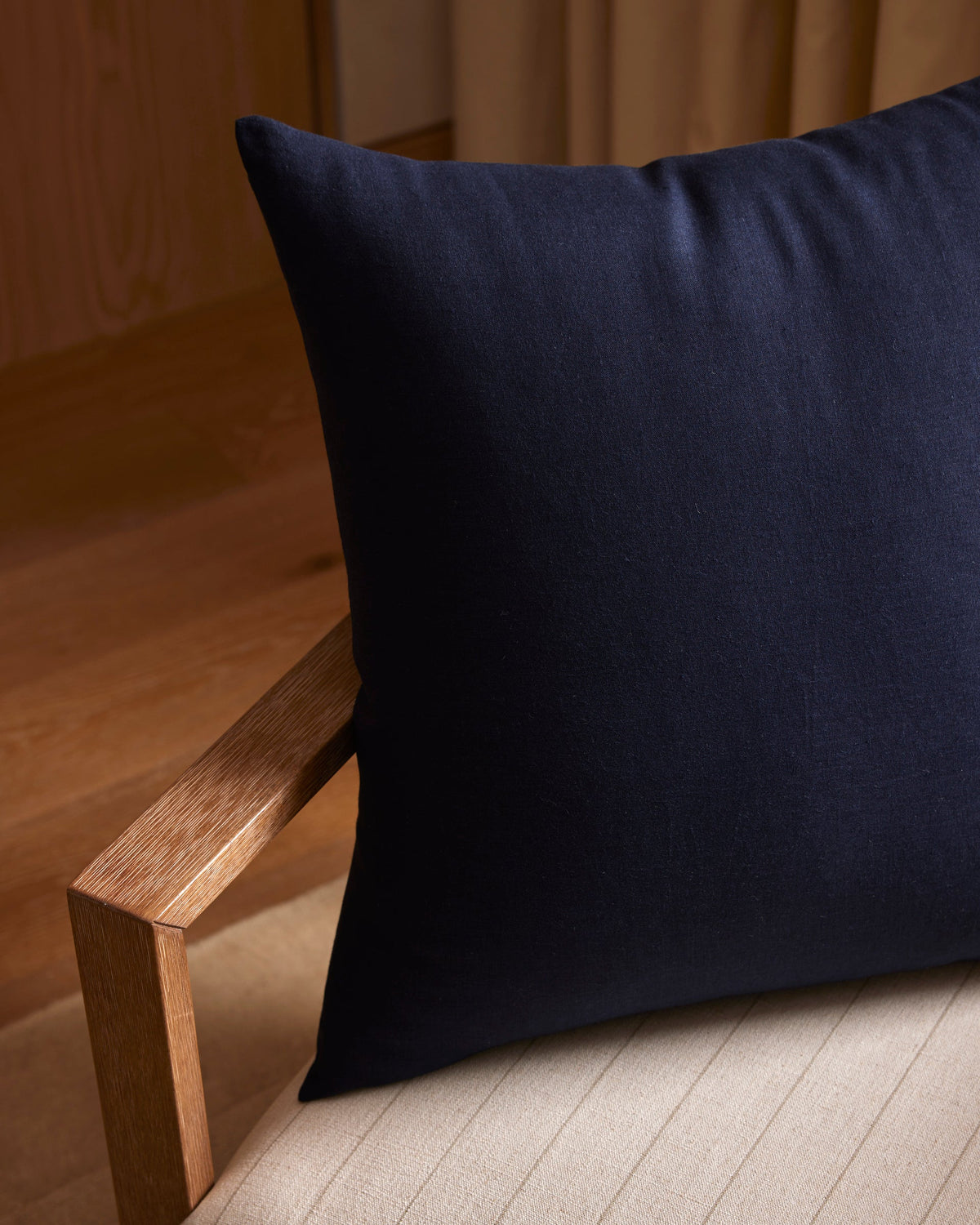 A close-up of a wooden chair with a light cushion and a large, plain dark navy blue Linen Throw Pillow by Cozy Earth resting against the backrest, set against wooden and beige surfaces.|Color:Eclipse|Size:26" x 26"