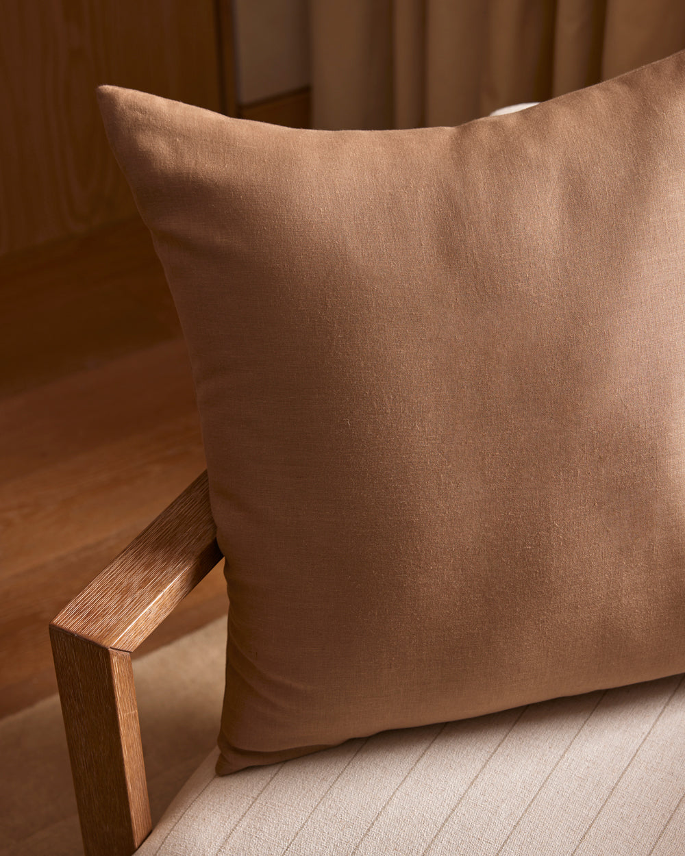 A Cozy Earth Linen Throw Pillow sits on a light beige upholstered chair with wooden armrests, surrounded by wood and neutral decor in a softly lit room.|Color:Maple|Size:26" x 26"