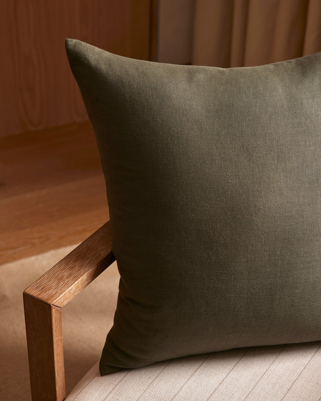 A Cozy Earth Linen Throw Pillow in dark green sits on a light beige cushioned chair with a wooden armrest, surrounded by warm lighting and wood and fabric accents in the background.|Color:Olive |Size:24" x 24"