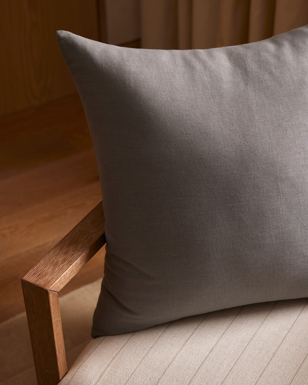 A Cozy Earth Linen Throw Pillow in gray sits on a light beige cushioned chair with a wooden armrest, set in a softly lit room featuring wooden floors and curtains in the background.|Color:Steel |Size:26" x 26"
